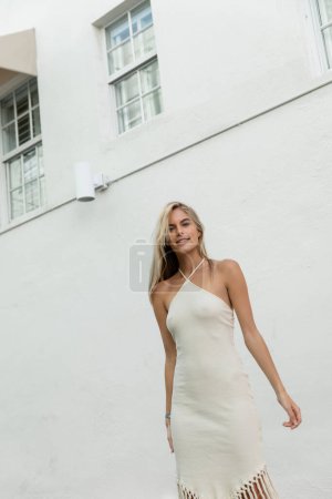 Eine junge blonde Frau in einem wallenden weißen Kleid posiert elegant vor einem prachtvollen Gebäude in Miami.
