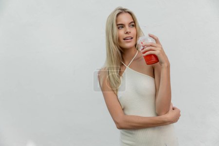 Eine junge, schöne blonde Frau in Miami hält eine rote Tasse in einem wallenden weißen Kleid unter der warmen Sonne.
