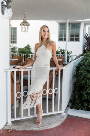 A young, blonde woman in a white dress stands gracefully on a balcony in Miami.