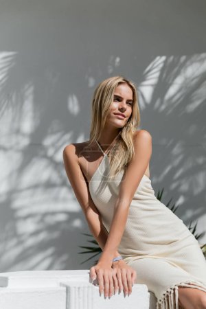 A young, beautiful blonde woman in a white dress leans against a wall in Miami, exuding serenity and elegance.