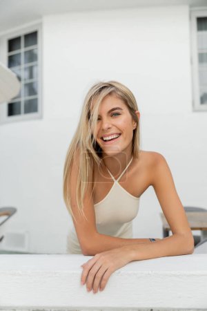 A young, beautiful blonde woman is elegantly leaning on a table in Miami, wearing a white tank top.