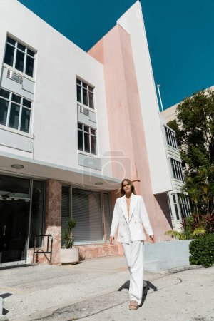 A young, beautiful blonde woman stands confidently in front of a striking building in Miami, exuding sophistication and style.