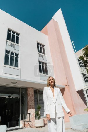 Eine junge, blonde Frau steht anmutig vor einem rosa-weißen Gebäude in Miami.