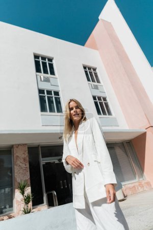 A young blonde woman stands gracefully in front of a striking pink and white building in Miami.