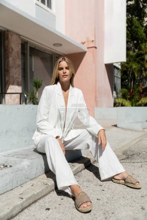 Eine abgeklärte junge Frau mit blonden Haaren, elegant gekleidet in einem weißen Anzug, sitzt anmutig auf dem Boden in Miami.