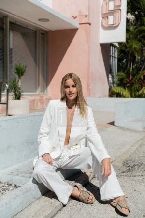 A young blonde woman in a chic white suit seated gracefully on the ground in a peaceful and contemplative pose.