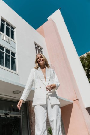 Una joven y hermosa mujer rubia se para con confianza en un traje blanco frente a un impresionante edificio de Miami.