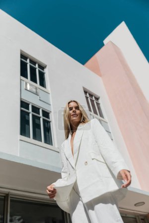 Foto de Una joven y hermosa rubia vestida con un traje blanco se para con confianza frente a un llamativo edificio urbano. - Imagen libre de derechos