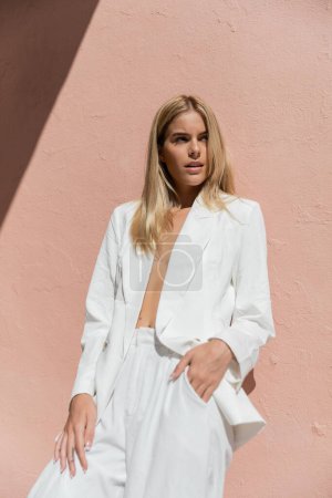 A blonde woman exudes elegance in a white suit, standing confidently in front of a vibrant pink wall.