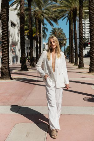 Foto de Una mujer rubia exuda confianza mientras camina por una calle de Miami en un impresionante traje blanco, una visión de elegancia. - Imagen libre de derechos