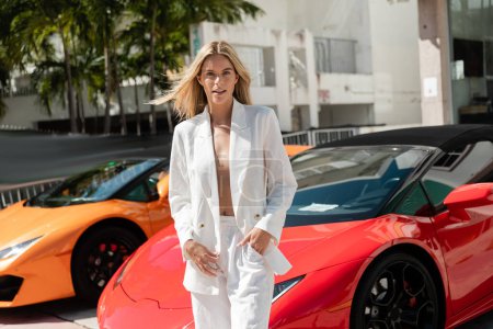 Una mujer rubia impresionante se para con gracia junto a un vibrante coche deportivo rojo en un glamuroso entorno de Miami.