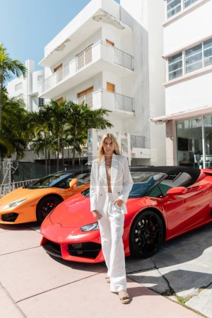Una joven rubia de pie con confianza frente a una fila de coches deportivos de lujo en Miami, exudando elegancia y sofisticación.