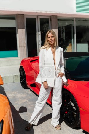 Eine junge, schöne blonde Frau steht selbstbewusst neben einem eleganten roten Sportwagen in Miami.