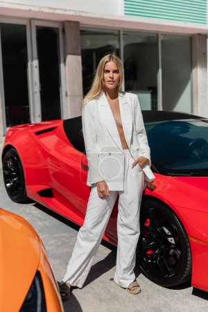 Eine junge, schöne blonde Frau steht selbstbewusst neben einem leuchtend roten Sportwagen im sonnigen Miami.