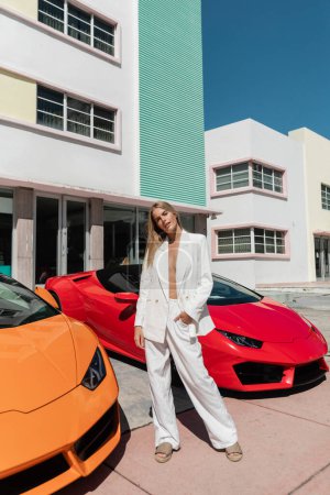 Una joven y hermosa rubia se para con confianza junto a dos elegantes coches deportivos en Miami.