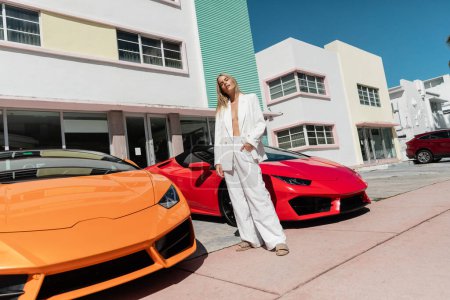 A young blonde woman stands confidently between two cars in front of a building.