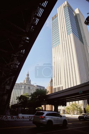 scenic view of skyscraper near car moving on roadway under bridge in new york city, urban atmosphere