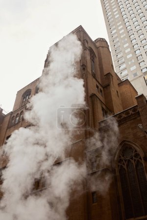 Dampf in der Nähe der katholischen Kirche und des Wolkenkratzers aus rotem Backstein an der urbanen Straße von New York City, niedriger Blickwinkel