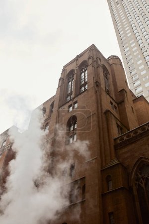 low angle view of red brick catholic church near steam and skyscraper on street in new york city