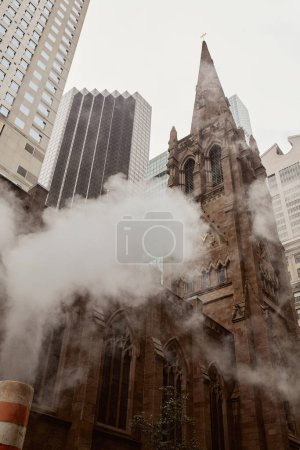 Blick auf die katholische Kirche aus rotem Backstein in der Nähe von Wolkenkratzern und Dampf auf der New Yorker City Street