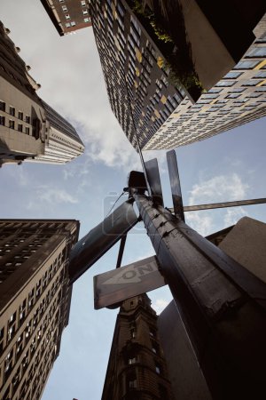 bottom view of street pole with traffic signs near tall buildings against blue sky in new york city