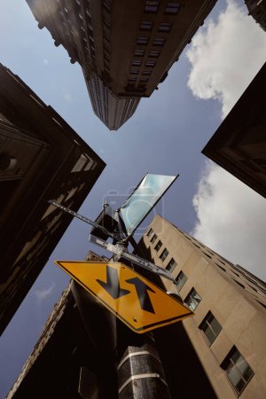 bottom view of street pole with traffic signs near tall buildings against blue sky in new york city