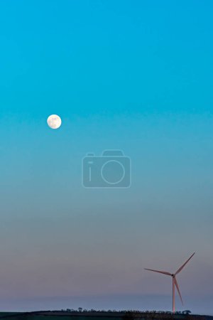 Ein atemberaubendes Porträt einer Windkraftanlage, die hoch vor dem Hintergrund eines lebhaften Sonnenuntergangs in Devon steht, bei dem der Mond anmutig aufgeht.