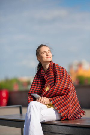Outdoor portrait of a 35-year-old woman sits on a bench covered with a blanket and basks in the spring sun. She holds a smartphone in her hands and communicates online.
