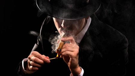 Foto de Close-up photo in retro style of a shaded detective in a black suit and hat lighting a cigar against a black background. - Imagen libre de derechos
