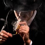 Close-up photo in retro style of a shaded detective in a black suit and hat lighting a cigar against a black background.