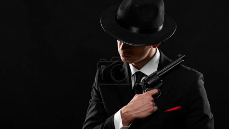 Un gangster des années 1940 armé. Homme en costume noir et chapeau avec un pistolet sur fond sombre. Photo avec espace de copie.