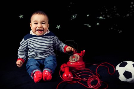 Photo for Portrait of smiling little boy wearing christmas hat near red vintage telephone sitting on black background - studio shot - Royalty Free Image
