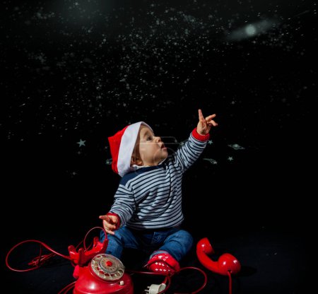 Photo for Portrait of smiling little boy wearing christmas hat near red vintage telephone sitting on black background - studio shot - Royalty Free Image