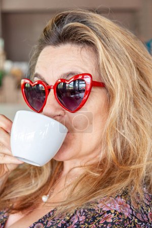 Téléchargez les photos : Belle femme d'âge moyen avec des lunettes en forme de coeur boire une tasse de café - en image libre de droit