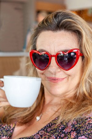 Photo for Beautiful middle aged woman with heart shaped glasses drinking a cup of coffee - Royalty Free Image