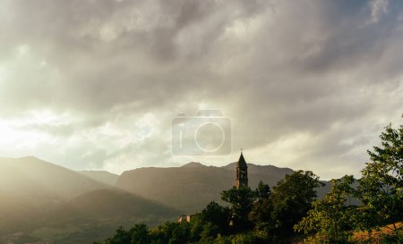 Téléchargez les photos : Ancien clocher dans un village médiéval entouré de collines au coucher du soleil avec le soleil obscurci par les nuages - en image libre de droit