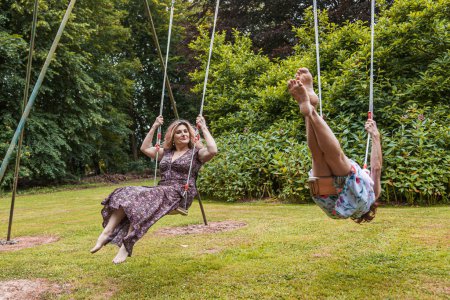 Téléchargez les photos : Beautiful middle aged couple of female friends swinging on children's swings in a french park - en image libre de droit