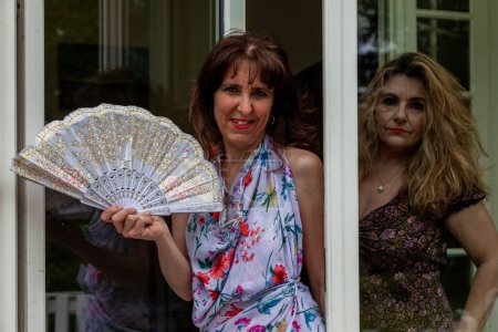 Téléchargez les photos : Pair of beautiful middle aged women waving a vintage fan in front of a french villa - en image libre de droit