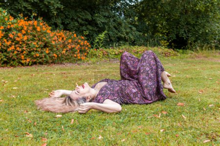 Téléchargez les photos : Pretty middle aged woman is laughing lying down on a lawn in a public park - en image libre de droit