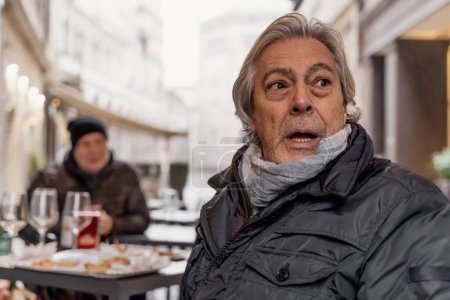 Foto de Portrait of mature man with surprised expression sitting outside a bar in winter - Imagen libre de derechos