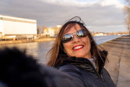 Téléchargez les photos : Femme d'âge moyen portant des vêtements d'hiver prendre un selfie par une rivière - concept de personnes dans les loisirs - en image libre de droit