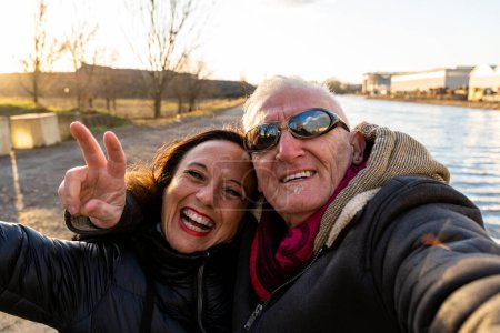 Téléchargez les photos : Couple d'âge moyen portant des vêtements d'hiver prendre un selfie sur les rives de la rivière - concept de personnes dans les loisirs - en image libre de droit
