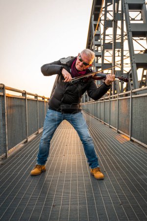 Téléchargez les photos : Attractive middle aged musician man playing an electric violin outdoors over an iron bridge - en image libre de droit