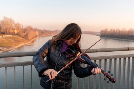 Photo for Attractive middle aged woman playing an electric violin outdoors over an iron bridge - Royalty Free Image