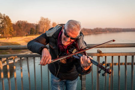 Photo for Attractive middle aged musician man showing an electric violin outdoors over an iron bridge - Royalty Free Image