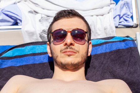 Foto de Portrait of young man wearing sunglasses and tanning in summer on a lounger - Imagen libre de derechos