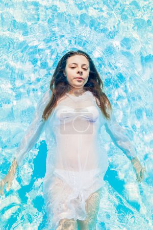 Téléchargez les photos : Jeune fille en maillot de bain flottant sur l'eau azur de la piscine à la lumière du soleil - Concept de belles personnes s'amusant en été - en image libre de droit