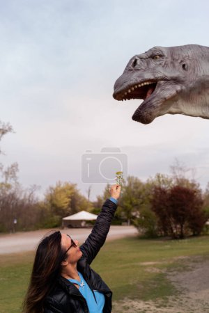 Photo for Middle aged tourist having fun feeding a flower to a life-size model dinosaur outdoors - Royalty Free Image