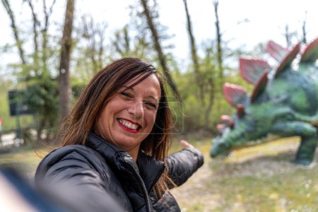Téléchargez les photos : Femme d'âge moyen souriante prenant un selfie devant des modèles de dinosaures dans un parc jurassique - concept de style de vie de vacances et de voyage - en image libre de droit