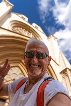 Foto de Guapo hombre de mediana edad visitando la catedral de Santa María, Tarragona - Feliz turista tomando un selfie frente a la iglesia - Concepto de turismo y vacaciones - Imagen libre de derechos